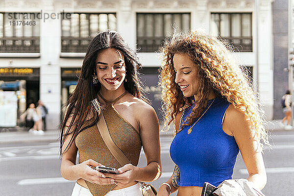Lächelnde Frau teilt ihr Mobiltelefon mit ihrer Freundin auf der Straße