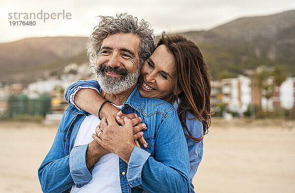 Glückliche ältere Frau umarmt Mann von hinten am Strand