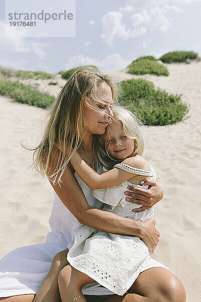 Lächelnde Tochter sitzt an einem sonnigen Tag auf dem Schoß der Mutter am Strand