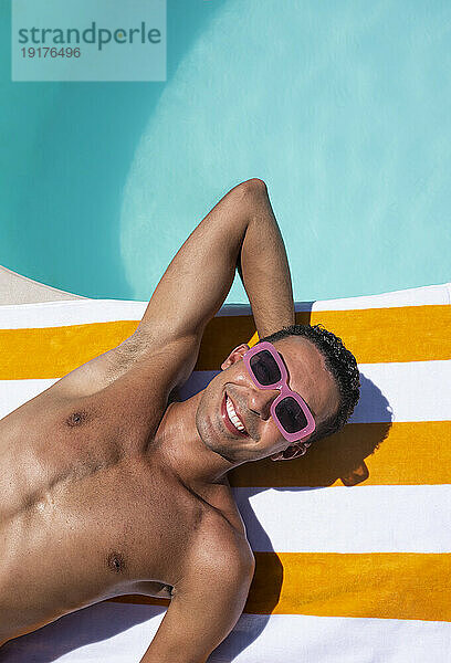 Lächelnder Mann mit Sonnenbrille entspannt sich auf einem Handtuch am Pool