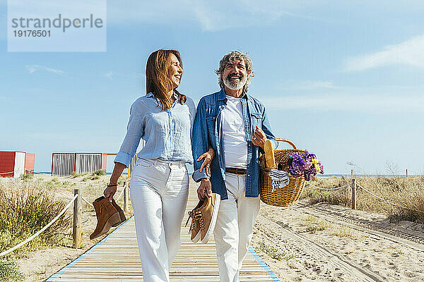 Happy senior woman and man carrying picnic basket and shoes walking at beach