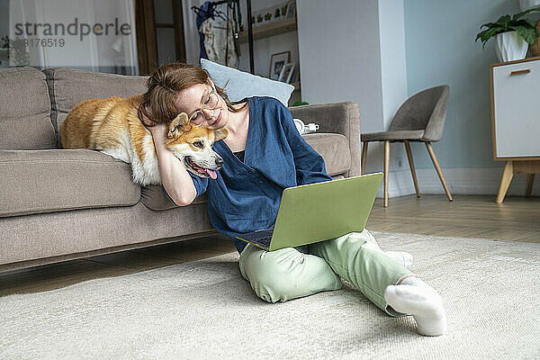 Frau benutzt Laptop und umarmt Hund im Wohnzimmer