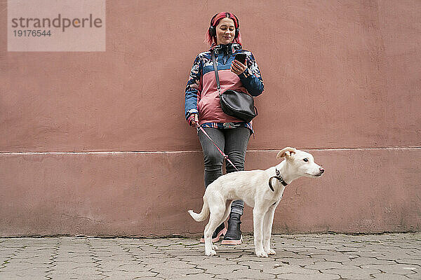 Lächelnde Frau steht mit Hund und benutzt Smartphone vor der Wand