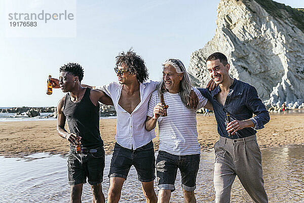 Multiethnische Freunde trinken Bier und spazieren an einem sonnigen Tag am Strand
