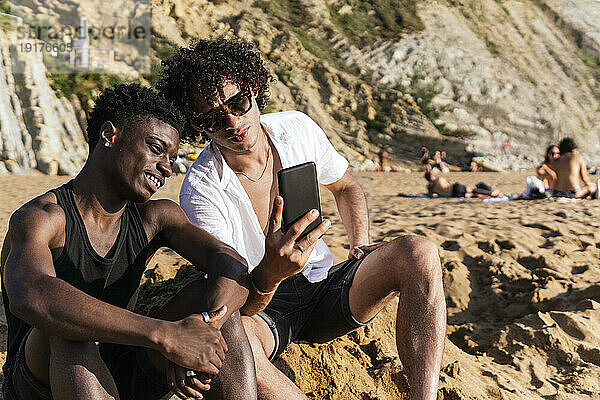 Junger Mann teilt Smartphone mit Freund  der auf Sand sitzt