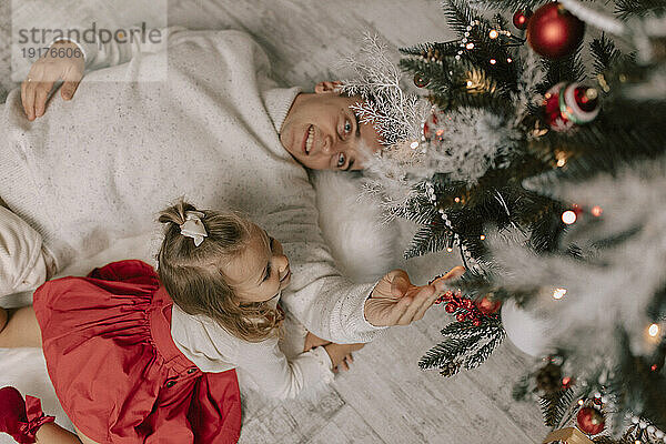 Verspielter Vater und Tochter liegen auf dem Boden und berühren den Weihnachtsbaum