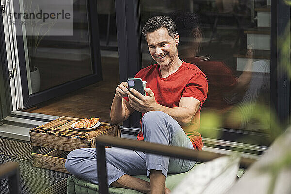 Mann entspannt sich auf Balkon mit Smartphone und Tasse in der Hand