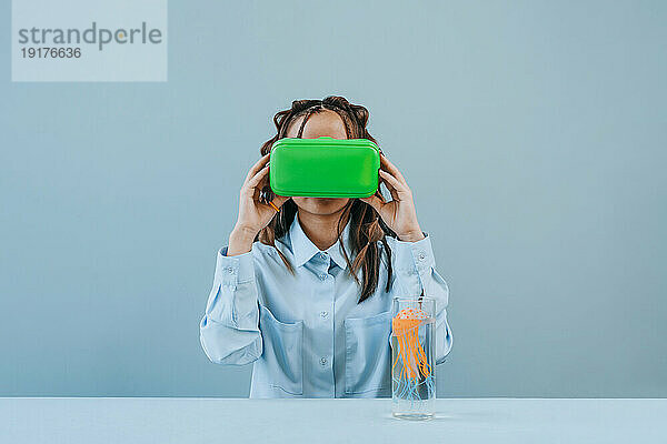 Junge Frau mit VR-Brille in der Nähe von Quallen im Trinkglas vor blauem Hintergrund