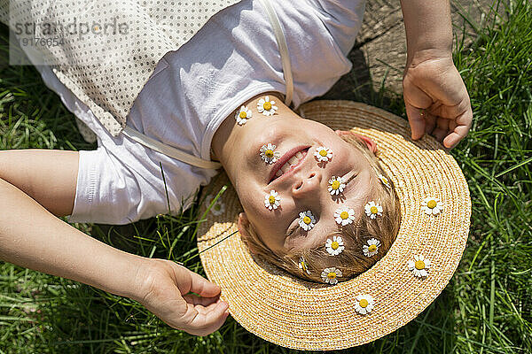 Fröhlicher Junge mit Gänseblümchen über dem Gesicht  der sich im Gras entspannt