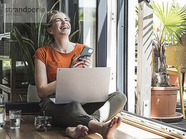 Portrait of woman sitting by open balcony laughing with smart phone in hands