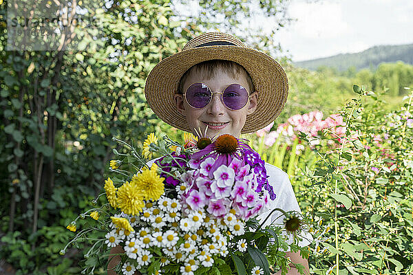 Fröhlicher Junge mit Hut und verschiedenen Blumen im Garten