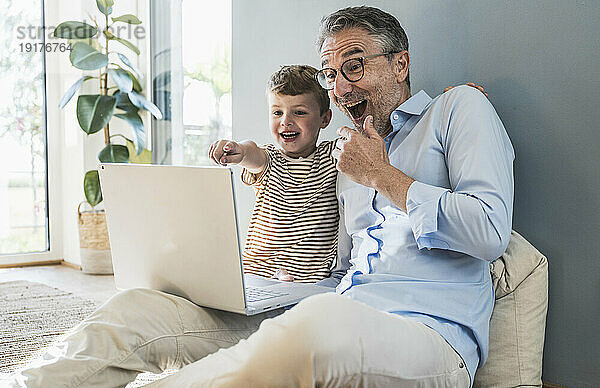 Fröhlicher Junge zeigt auf Laptop  der neben Großvater im Wohnzimmer sitzt