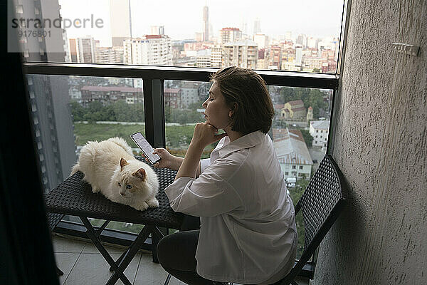 Frau sitzt mit Katze und benutzt Smartphone auf Balkon