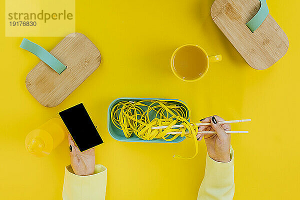 Hände einer Frau mit kabelgebundenen Nudeln und Mobiltelefon auf gelbem Tisch