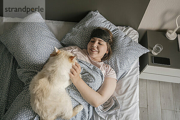 Lächelnde Frau mit Augenmaske streichelt Katze auf dem Bett