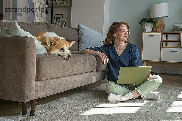 Junger Freiberufler mit Laptop sitzt neben seinem Hund im Wohnzimmer