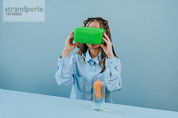 Frau beobachtet VR-Brille mit Quallen im Glas auf dem Tisch vor blauem Hintergrund