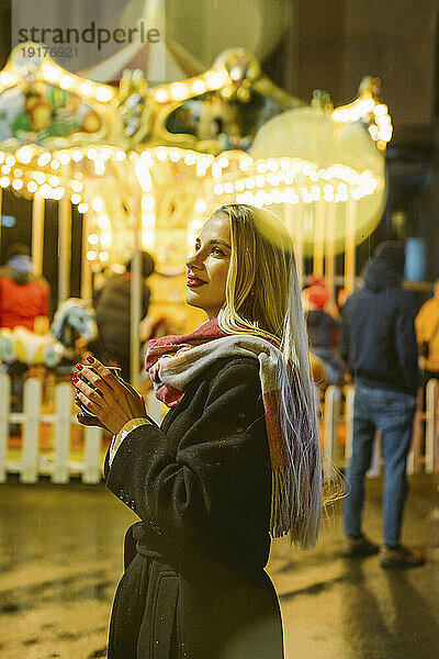 Lächelnde blonde Frau mit langen Haaren hält eine Tasse Glühwein in der Hand und steht auf dem Weihnachtsmarkt