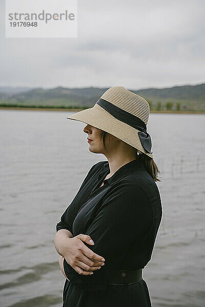 Woman wearing straw hat with arms crossed near lake