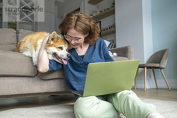 Lächelnder Freiberufler mit Laptop umarmt Hund im Wohnzimmer