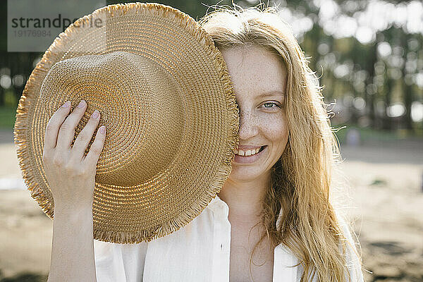 Blonde Frau versteckt ihr Gesicht mit Sonnenhut am Strand