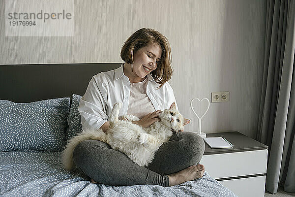 Smiling woman sitting cross legged and stroking cat on bed at home