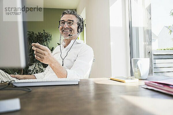 Lächelnder Freiberufler  der am Schreibtisch im Heimbüro sitzt und über ein Headset spricht