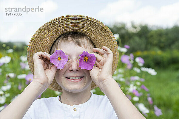 Glücklicher Junge hält rosa Blumen über den Augen im Garten