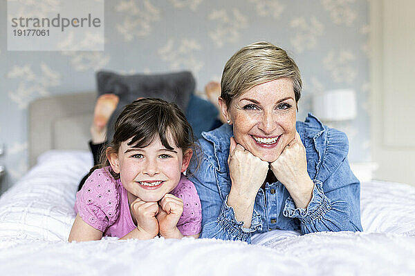 Happy mother and daughter with hands on chins lying in bedroom