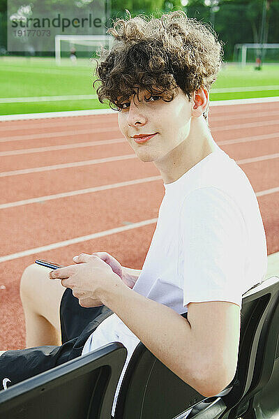 Lächelnder Teenager mit Smartphone sitzt im Stadion