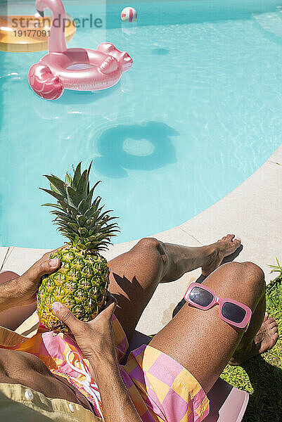 Junger Mann sitzt mit Ananas und Sonnenbrille am Pool
