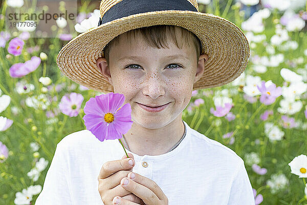 Lächelnder Junge mit Hut hält Blume im Garten