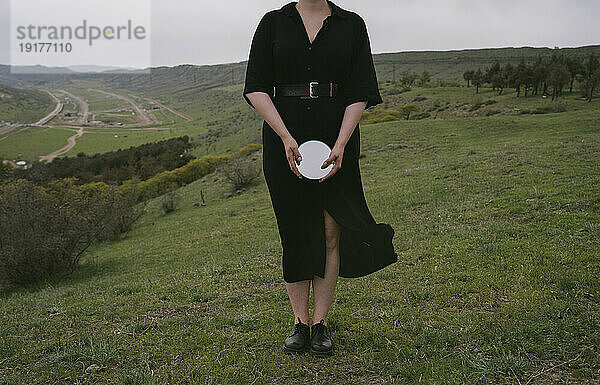 Frau im schwarzen Kleid hält Spiegel vor grüner Landschaft