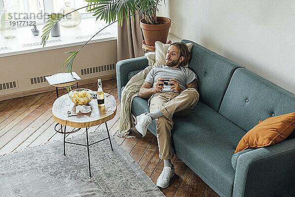 Mann mit Gamecontroller liegt zu Hause auf dem Sofa