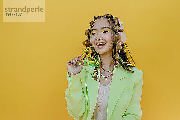 Happy woman with headphones holding sunglasses against yellow background