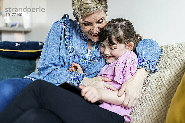 Happy mother embracing daughter holding tablet PC and sitting on sofa