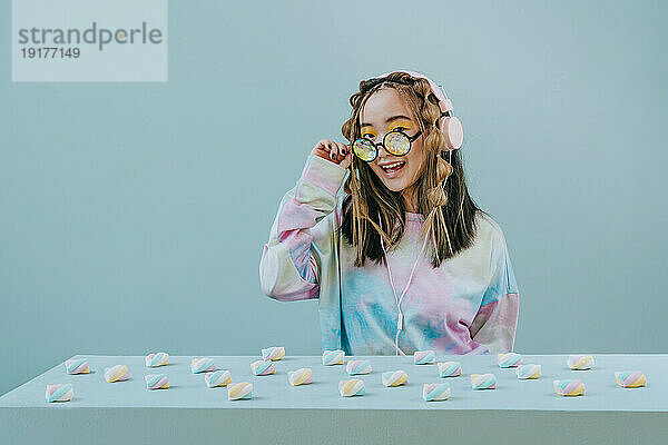 Happy young with marshmallows on table wearing kaleidoscope glasses against blue background