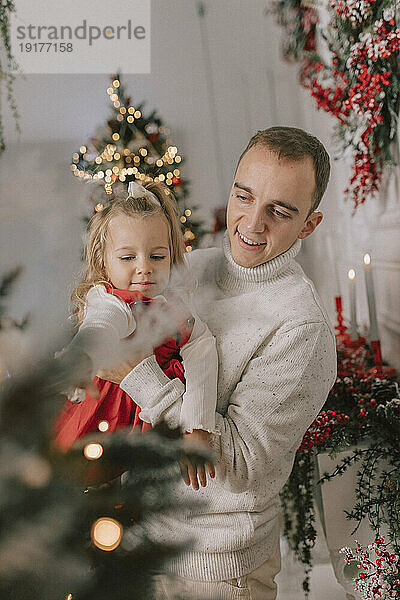Glücklicher Vater trägt seine Tochter und schaut auf den Weihnachtsbaum