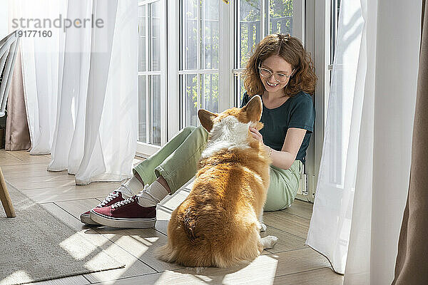 Smiling woman caressing Welsh Corgi dog sitting at home