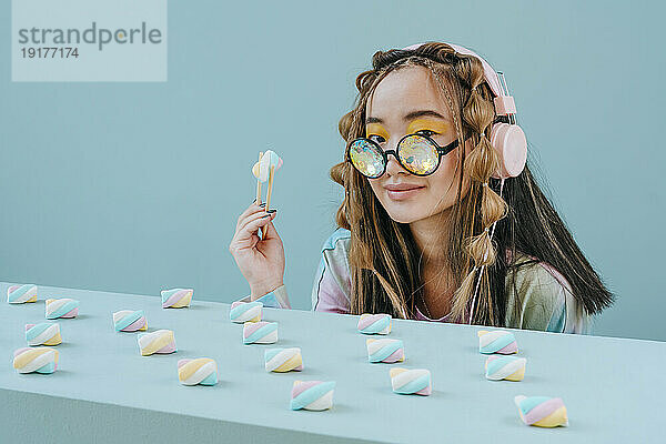 Junge Frau mit Kopfhörern und Kaleidoskopbrille isst Marshmallow im Studio