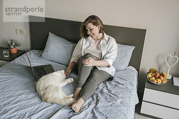 Lächelnde Frau spielt zu Hause mit Katze neben Laptop im Bett