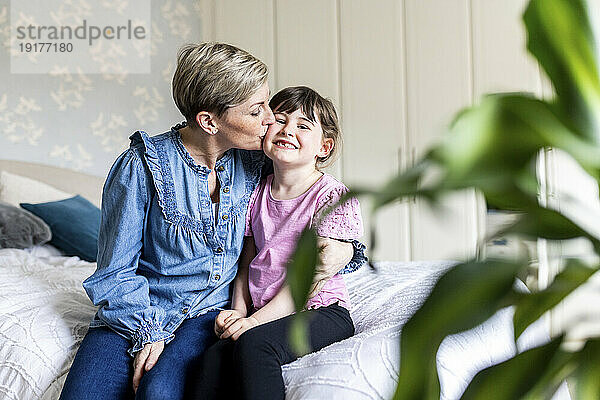 Mature woman kissing daughter sitting on bed at home