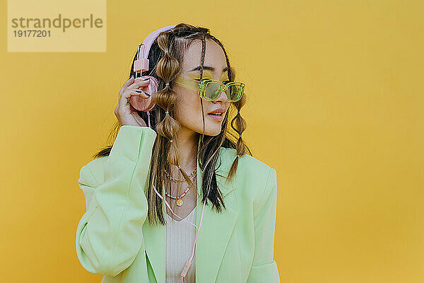 Junge Frau mit Sonnenbrille hört Musik über Kopfhörer vor gelbem Hintergrund
