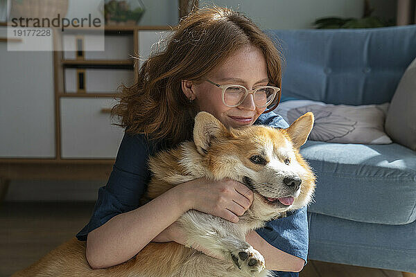 Lächelnde Frau umarmt walisischen Corgi-Hund im Wohnzimmer