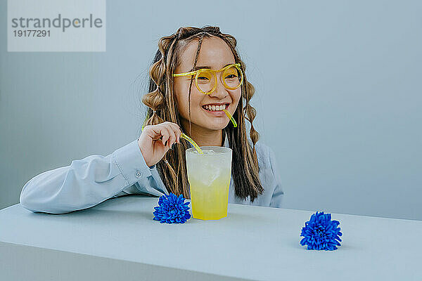 Lächelnde junge Frau trinkt mit glasartigem Strohhalm am Tisch im Studio