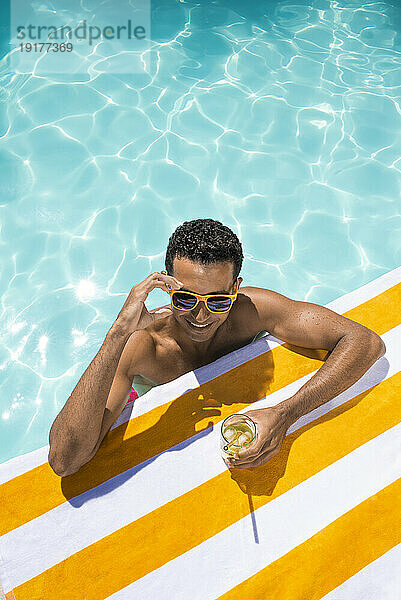 Glücklicher junger Mann mit Getränk  der sich am Pool auf ein Handtuch stützt
