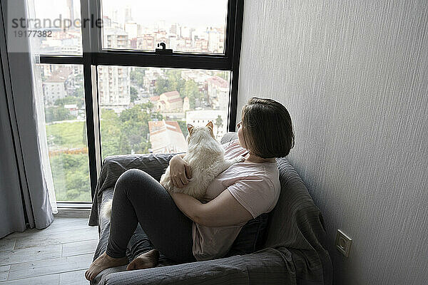 Nachdenkliche junge Frau sitzt mit Katze zu Hause auf dem Sofa