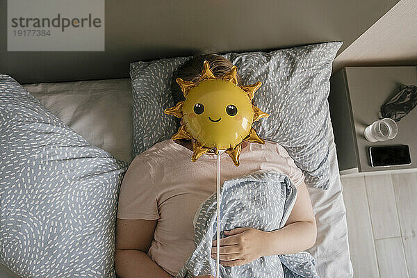 Woman hiding face with balloon on bed at home