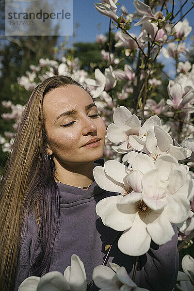 Frau mit geschlossenen Augen durch Magnolienblüte im Park an einem sonnigen Tag