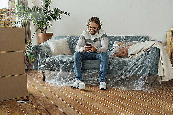 Lächelnder junger Mann  der zu Hause auf dem Sofa sein Mobiltelefon benutzt